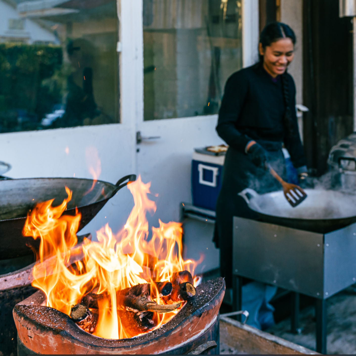 Köchin Gin Eymann steht an ihrer Outdoor-Küche und rührt im Topf. Im Vordergrund brennt ein Feuer im traditionellen Holzkohleofen. 